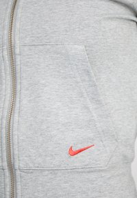 Hanorac Nike cu fermoar, de culoare gri deschis, cu un material texturat, buzunare frontale și un mic logo Nike roșu.