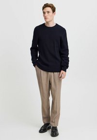 Pulover bleu în dungi cu guler rotund, asociat cu pantaloni bej croiți și pantofi negri din piele. Modelul stă cu mâinile în buzunare.
