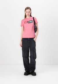 Camiseta rosa con logotipo, pantalones cargo oscuros con bolsillos y zapatos de plataforma negros. La modelo sostiene una pequeña bolsa negra. Look simple y casual.