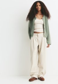 Frau mit langen lockigen Haaren, die eine grüne Strickjacke, ein beiges Tanktop, weite beige Hosen und braune Schnürschuhe trägt, steht an einer weißen Wand.