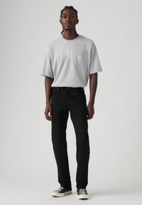 Muškarac u sivoj oversized majici s džepom na prsima, crnim uskim trapericama i crnim niskim tenisicama stoji naspram jednostavne pozadine.