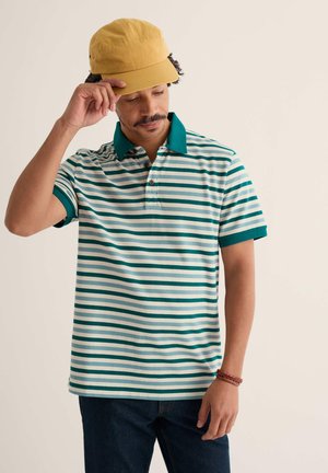 Uomo con cappellino giallo, polo a righe turchese e bianche, jeans scuri e braccialetto marrone intrecciato, che si sistema il cappellino con la mano.