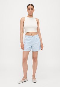 Shorts rayés bleu clair en coton, avec fermeture à bouton et poches latérales, associés à un débardeur blanc court et des chaussures plates.