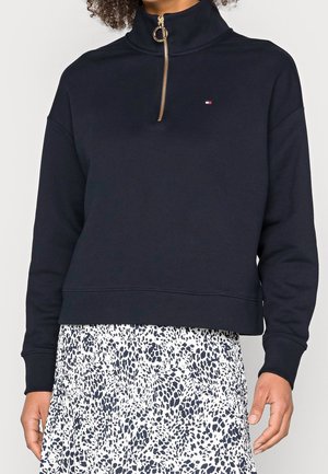 Femme portant un pull noir avec une demi-fermeture éclair dorée et un petit logo, associé à une jupe blanche à motifs noirs tachetés.