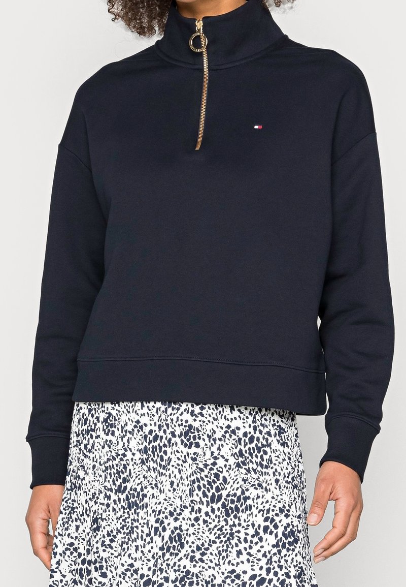 Femme portant un pull noir avec une demi-fermeture éclair dorée et un petit logo, associé à une jupe blanche à motifs noirs tachetés.