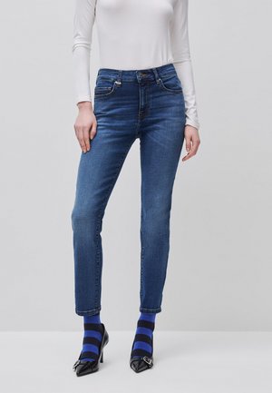 Frau trägt eng anliegende blaue Jeans, weißes langärmeliges Oberteil, schwarze High Heels und blau-schwarz gestreifte Socken und steht vor einem grauen Hintergrund.