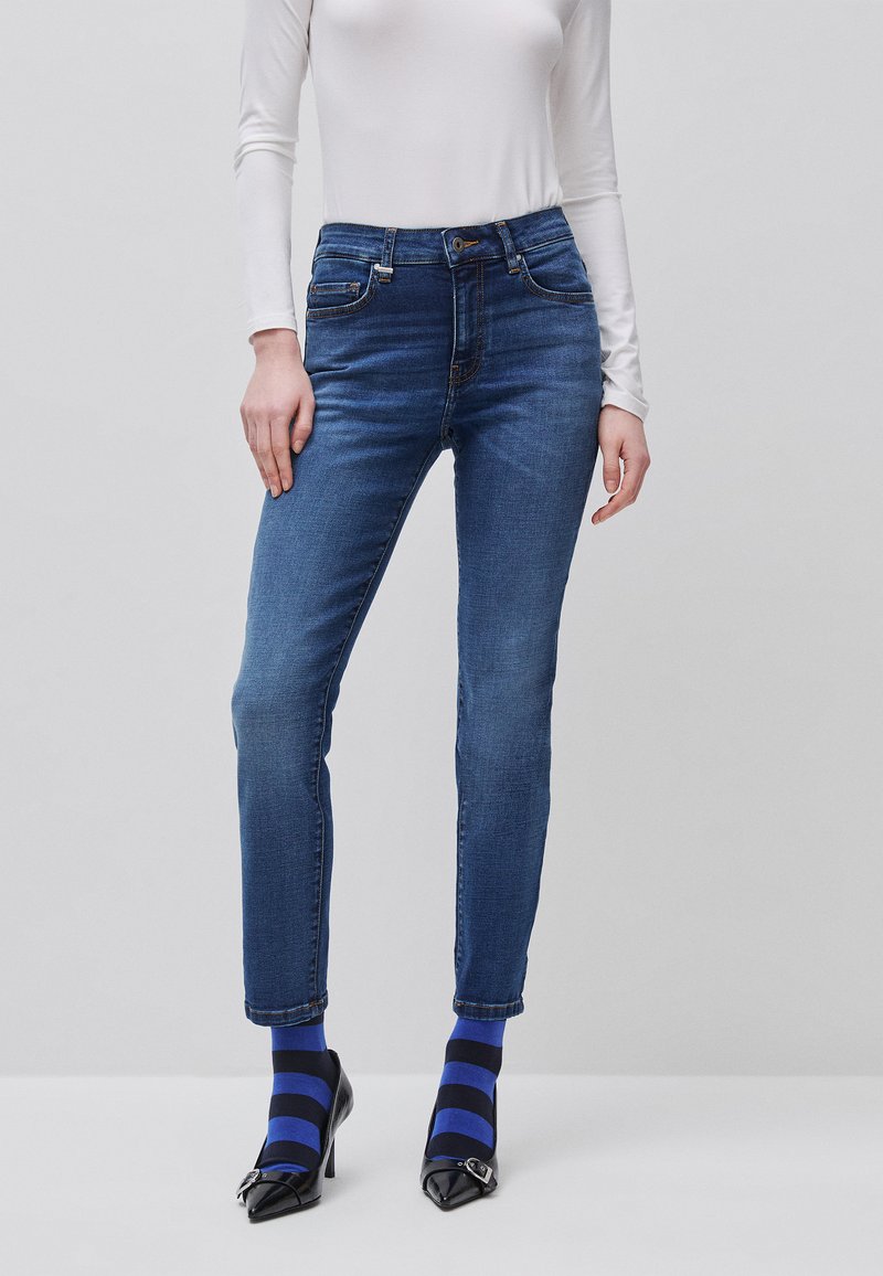 Vrouw draagt nauwsluitende blauwe jeans, wit langärmelig shirt, zwarte hoge hakken en blauw-zwarte gestreepte sokken, staand tegen een grijze achtergrond.