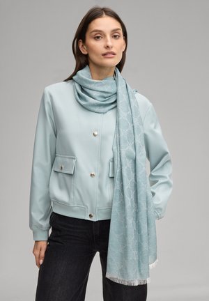 Vrouw draagt een lichtblauwe jas met gouden knopen en een blauw sjaal met patroon over haar schouder, staand tegen een effen grijze achtergrond.