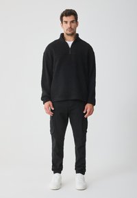 Pullover in fleece nero con zip a un quarto, caratterizzato da una consistenza morbida. Abbinato a pantaloni cargo neri e sneakers bianche. Design semplice.