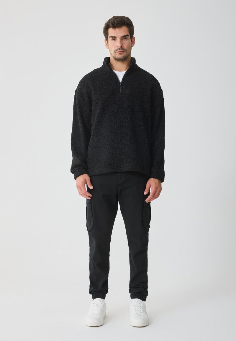 Pullover in fleece nero con zip a un quarto, caratterizzato da una consistenza morbida. Abbinato a pantaloni cargo neri e sneakers bianche. Design semplice.