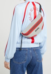 Bolso de cintura gris con acentos rojos, con un acabado suave, compartimento con cremallera y la palabra "KARL" impresa en la parte frontal.