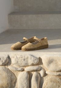 Beżowe espadryle zamszowe z zakrytym noskiem, paskiem wokół kostki i splecioną juty podeszwą, ustawione na teksturowanej kamiennej powierzchni.