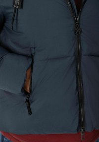 Dunkelblaue Daunenjacke mit glatter, strukturierter Stoffoberfläche, Reißverschluss vorne und Seitentasche. Auffällige Reißverschlussbeschläge sind schwarz.