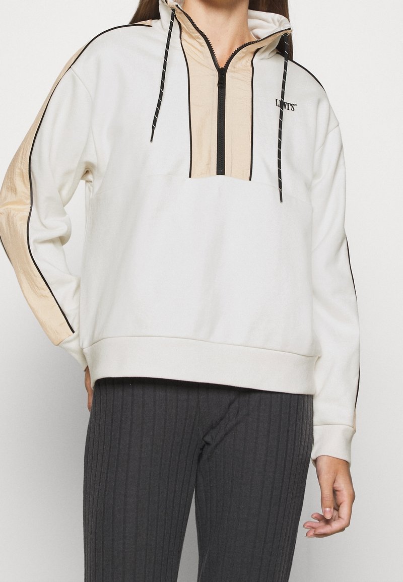 Vit huva-pullover med halv zip framtill, kontrasterande beige panel, svarta dragsnören och svarta detaljer. Textlogo på bröstet.