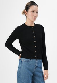 Jovem mulher com o cabelo preso, vestindo um cardigan preto com botões e jeans azuis, de pé com uma mão no bolso, contra um fundo branco.