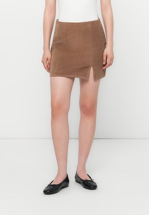 Vrouw die een bruine corduroy minirok met een voorste split draagt, gecombineerd met zwarte balletschoentjes en een wit topje, staand tegen een witte achtergrond.