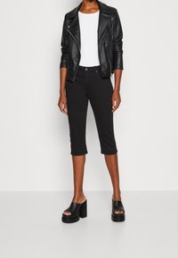 Osoba v černých zkrácených kalhotách, černé kožené bundě, bílém topu a černých platformových sandálech na podpatku stojí na jednobarevném pozadí.