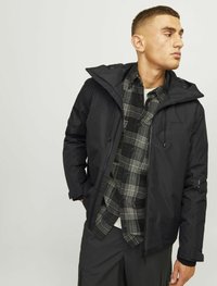 Chaqueta negra con capucha y cordones ajustables, que presenta una textura suave y un cierre de cremallera, superpuesta a una camisa de cuadros en blanco y negro.