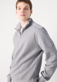 Sweat à fermeture éclair gris avec un design texturé, col côtelé et accents d'épaule. Présente une patch logo sur la manche gauche.