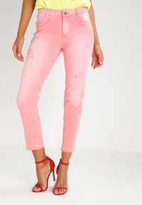 Jeans rosa con un diseño desvanecido, que presenta parches desgastados y bordados florales. Combinados con sandalias de tacón rojas.