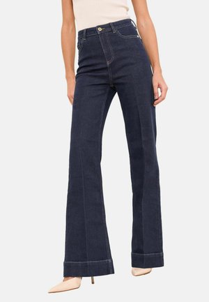 Tmavomodré džínsové zvonové nohavice s vysokým pásom, jemnými prešívacími detailmi a širokým, ohnutým lemom. Kombinované so svetlými lodičkami.