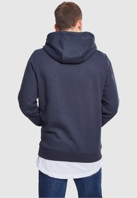 Laivastonsininen huppari, jossa on pehmeä tekstuuri, suuri huppu, ribbineulottu hihansuut ja suora helma. P äällä valkoinen kerrospaita.