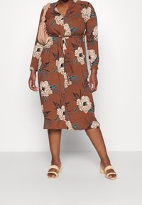 Kvinde iført en brun blomstret midikjole med lange ærmer, bærende på en blush skuldertaske og beige hæle sandaler.