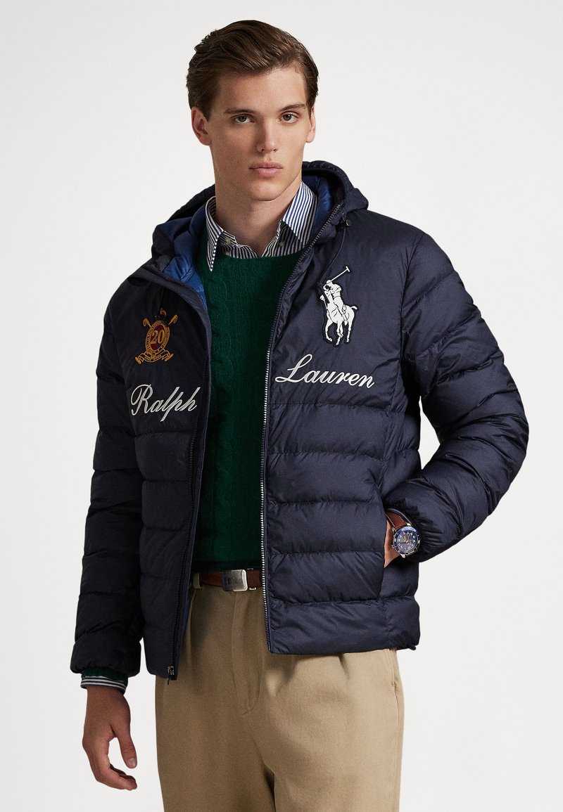 Jauns vīrietis, valkājot tumši zilu Ralph Lauren pūkaino jaku pār zaļu džemperi un svītrainu kreklu, ar bēšām biksēm un rokas pulksteni, stāvot.