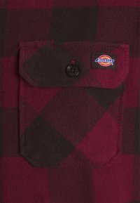 Poche en tissu à carreaux rouge foncé et noir avec un bouton noir et une petite étiquette multicolore de la marque Dickies sur le rabat.