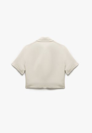 Przycięta, krótko rękawowa koszula w jasnobeżowym materiale o gładkiej fakturze i subtelnym detalu kołnierza; brak widocznych zapięć lub wzorów.