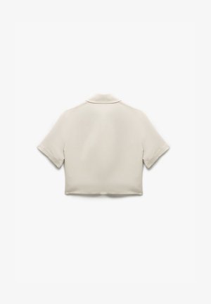 Przycięta, krótko rękawowa koszula w jasnobeżowym materiale o gładkiej fakturze i subtelnym detalu kołnierza; brak widocznych zapięć lub wzorów.