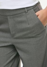 Šedé kalhoty s herringbone vzorem, hladkým povrchem, rovnými záhyby, poutky na pás a bočními kapsami, částečně viditelné v detailu.