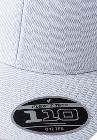 Lys grå baseballcap lavet af struktureret stof med en rund skygge. Har et sort cirkulært mærke med "FLEXFIT TECH 110 ONE TEN."