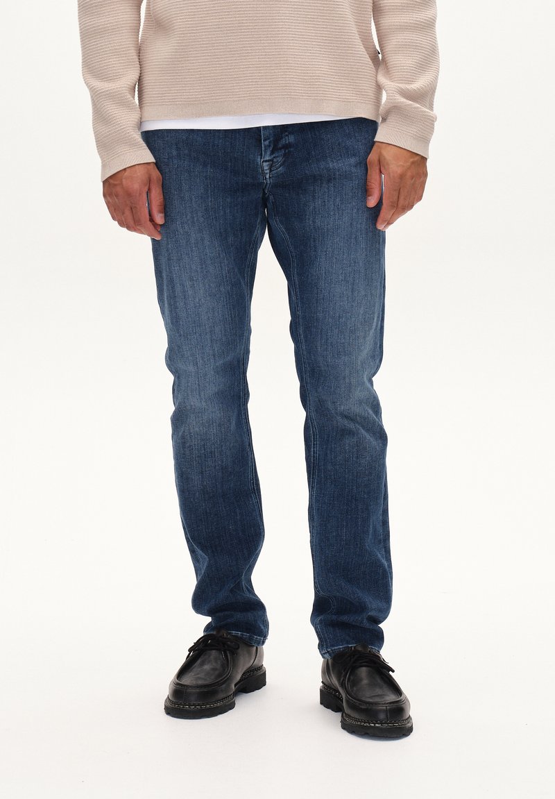 Temno modri denim kavbojki s ravnim krojem, ki vključujejo subtilne gube, rahlo bledenje in klasičen pet žepni dizajn. Obute z črnimi čevlji.