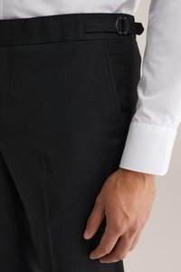 Schwarze maßgeschneiderte Hose mit flachem Vorderteil, Seitentaschen und einer Schnallendetails am Bund, kombiniert mit einem weißen Hemdärmel.