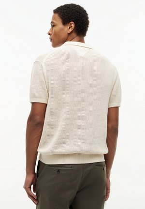 Άνδρας που φοράει ένα κρεμ πλεκτό πόλο με κοντά μανίκια και σκούρο πράσινο παντελόνι, με την πλάτη στραμμένη, σε απλό λευκό φόντο.