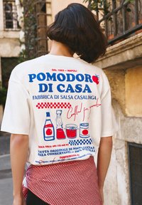 Personne aux cheveux courts et foncés portant une chemise blanche avec des motifs de sauce tomate italienne et un tissu rouge à carreaux noué autour de la taille.
