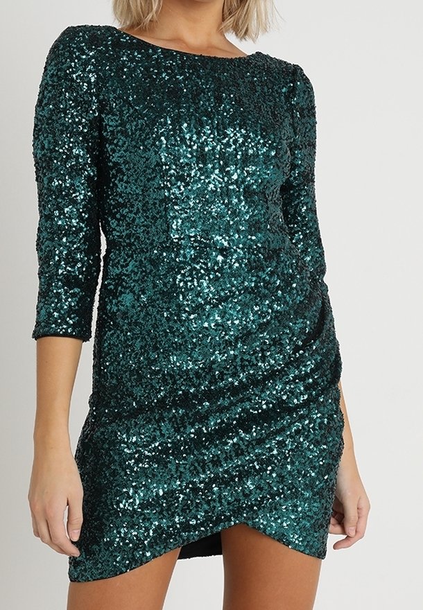 Femme portant une robe ajustée vert foncé à paillettes, avec des manches trois-quarts et un ourlet asymétrique.