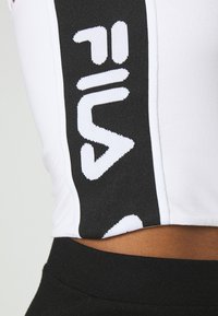 Biela crop top s čiernym bočným panelom a veľkým bielym logom "FILA", so žebrovanou textúrou, spárovaná s čiernymi legínami s vysokým pásom.
