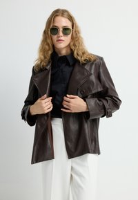 Giacca di pelle marrone con rever a punta, tasche frontali e dettagli con bottoni, abbinata a una camicia nera e pantaloni bianchi.