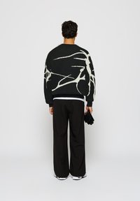 Hombre de pie de espaldas, vistiendo un suéter de patrón abstracto en blanco y negro, pantalones anchos negros, zapatillas blancas, sosteniendo guantes negros.