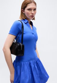 Žena s krátkymi vlasmi, ktorá má na sebe modré šaty s golierom a strieborné náušnice, nesie malú čiernu kabelku na ľavom ramene.