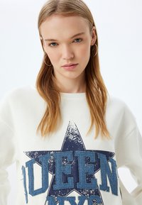 Sudadera blanca con un gran estampado de estrella azul desgastada y la palabra "QUEEN" en denim texturizado. Diseño de mangas largas y cuello redondo.