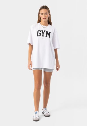 Bílá oversized tričko s výrazným černým potiskem "GYM". V kombinaci se šedými síťovanými šortkami a bílými teniskami s černými detaily.