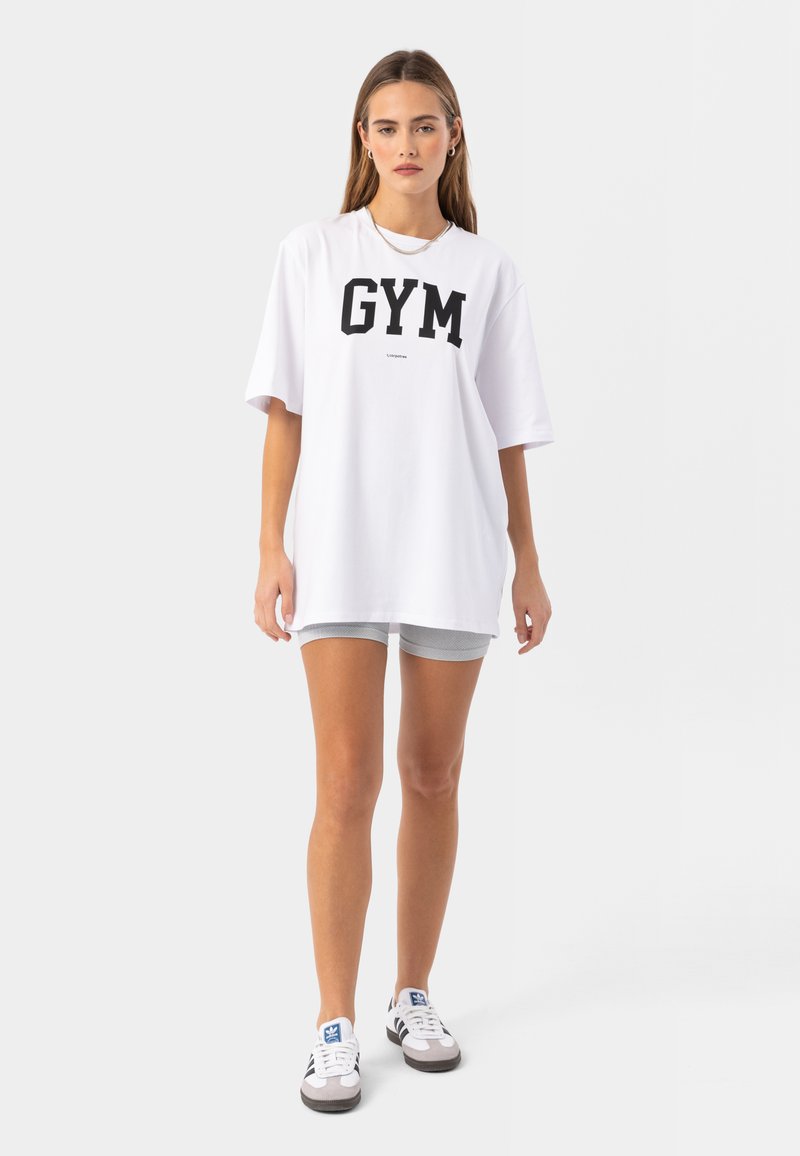 Biela oversized tričko s výrazným čiernym potlačou "GYM". V kombinácii so šedými sieťovanými šortkami a bielymi teniskami s čiernymi akcentmi.