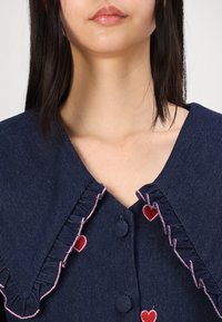 Σκούρο τζιν ύφασμα με φουσκωτό γιακά, ροζ ραφή και κεντημένη κόκκινη καρδιά. Διαθέτει δύο κουμπιά στο κέντρο.