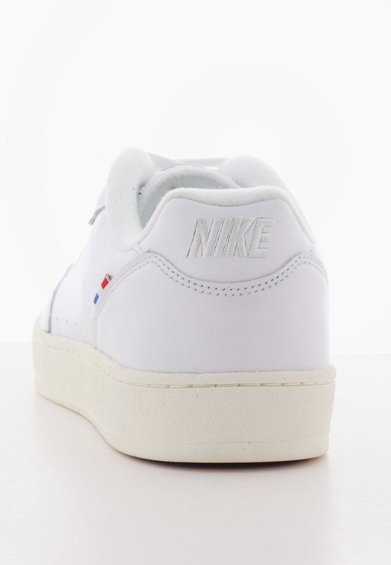 Weiße Leder-Sneaker mit gepolstertem Kragen, strukturiertem Gummisohle und gesticktem Nike-Logo. Mit kleinen blauen und roten Akzenten.