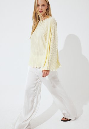 Blonde vrouw die loopt, gekleed in een lichtgele geplooide top met lange mouwen, brede witte broek en zwarte sandalen, tegen een witte achtergrond.