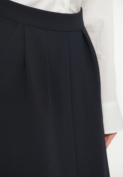 Σατέν φούστα ναυτικού χρώματος με πτυχώσεις, συνδυασμένη με λευκή πουκαμίσα με μακριά μανίκια, με τον καρπό να είναι ορατός από τη δεξιά πλευρά.