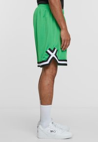 Groene basketbalschoenen met een mesh-textuur, voorzien van zwart-witte accenten aan de zoom en een 'X'-ontwerp. Gemaakt in combinatie met witte sneakers.